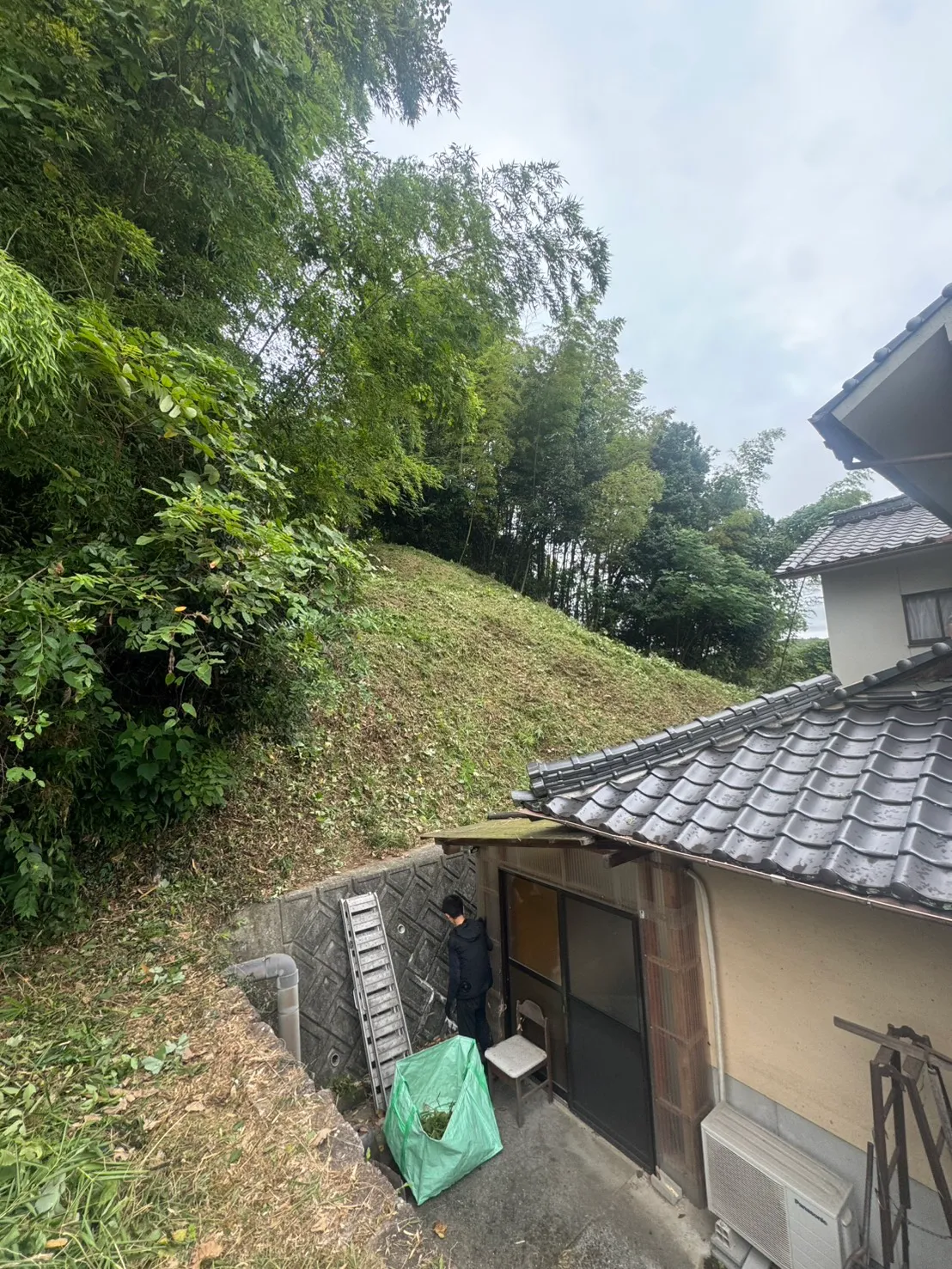 瓦屋根と擁壁の背後に刈り跡のある斜面と作業者