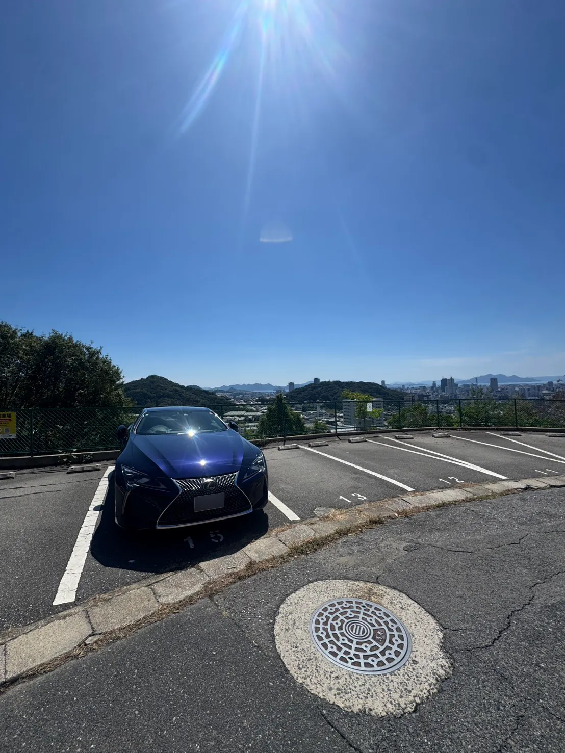 牛田早稲田の伐採後。同じ駐車区画付近でフェンス越しの高い枝葉が減り、奥の街並みと山並みが見える。