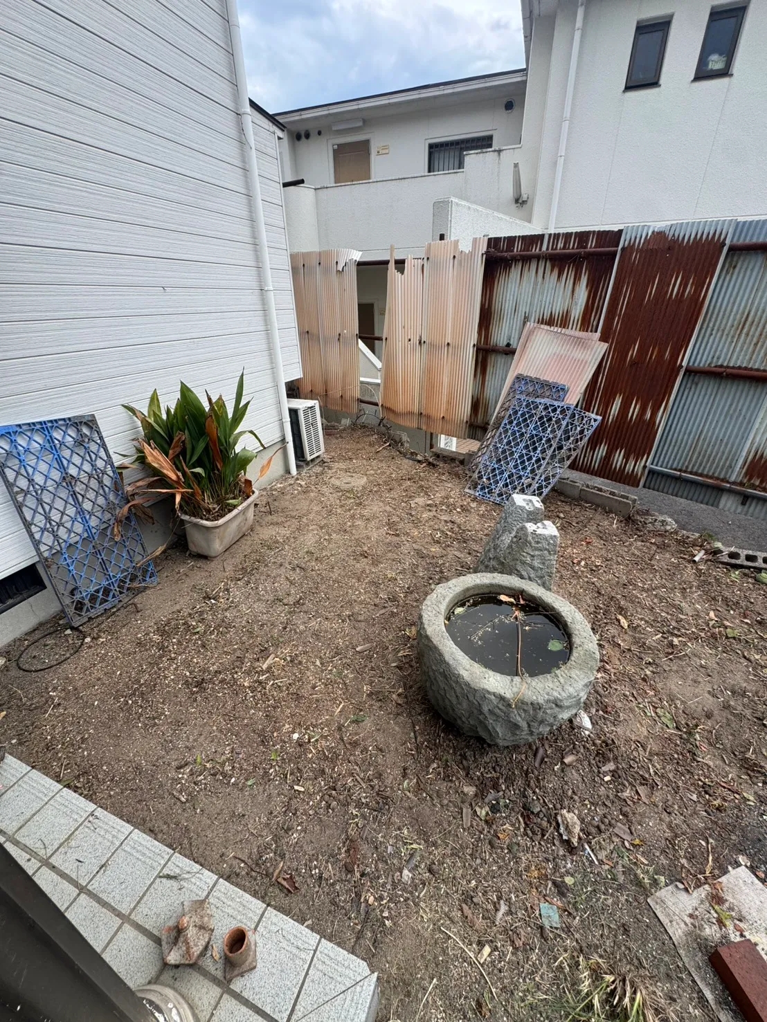 石の水鉢と鉢植えがある庭の一角。建物際から囲い沿いまで地面が見える