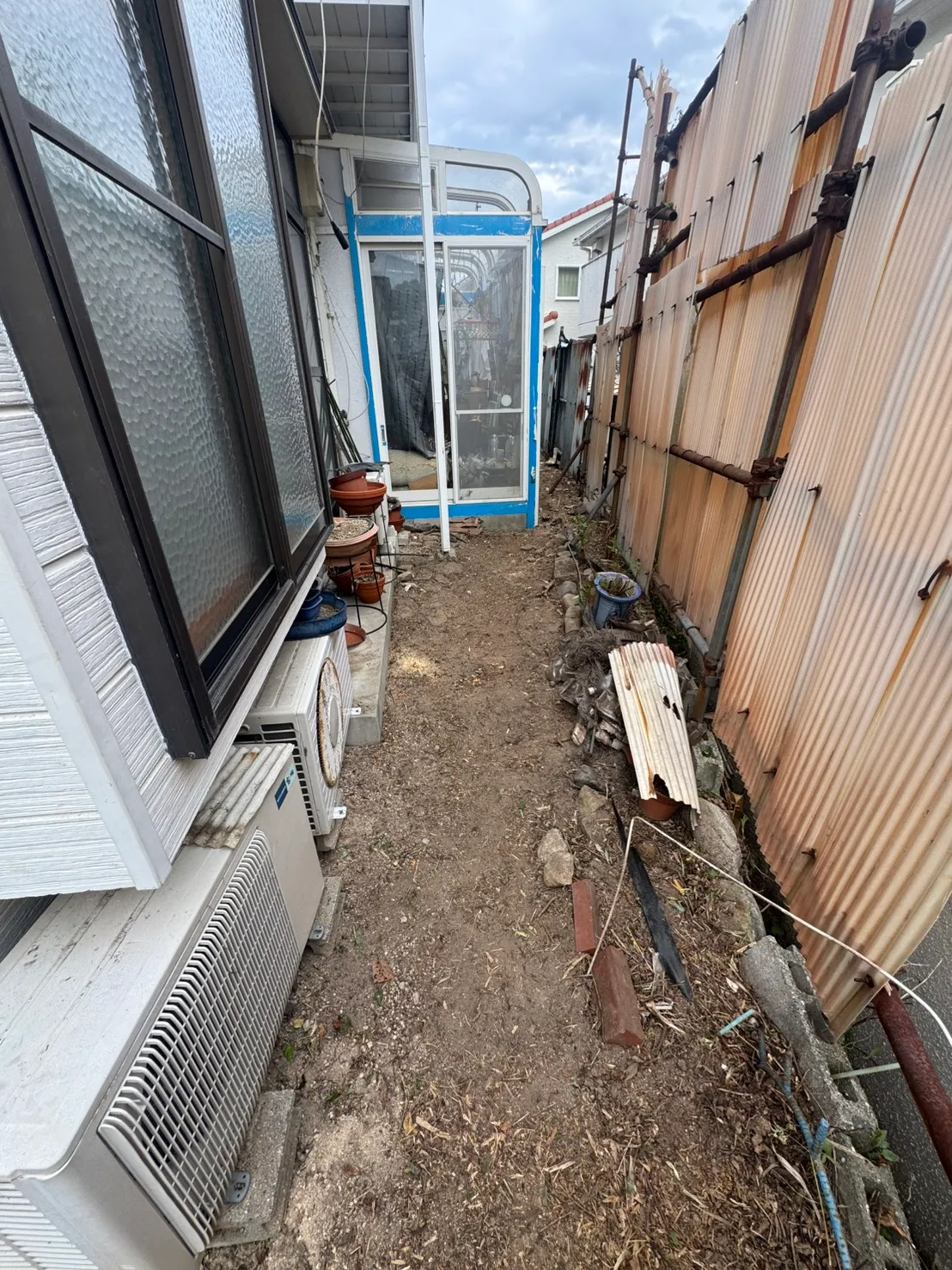 住宅の外壁と波板の囲いの間にある細い場所。室外機の横から奥へ地面が続いている
