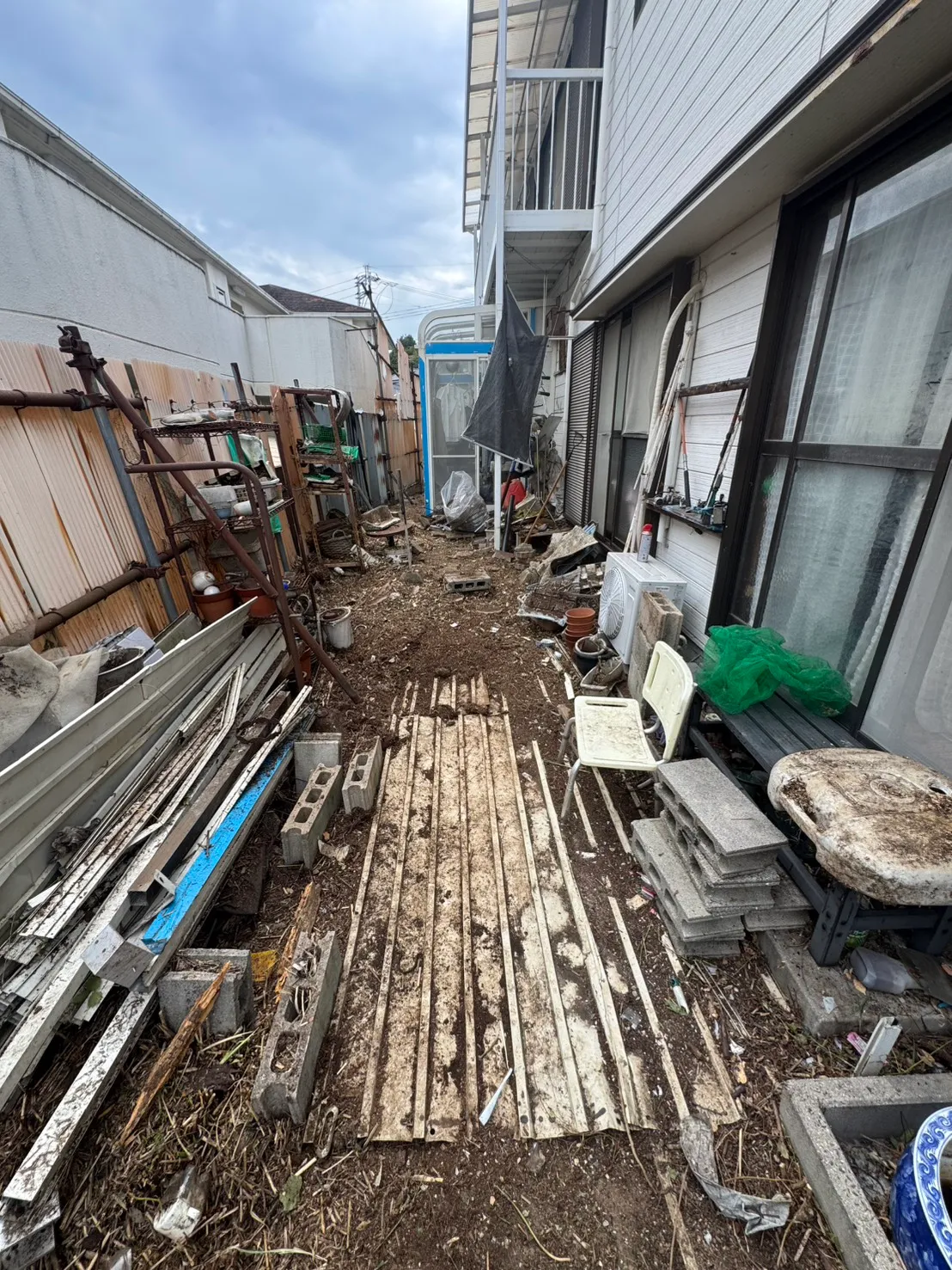 住宅外壁と囲いの間に鉢や資材が置かれ、その間の地面が見える施工後の状態