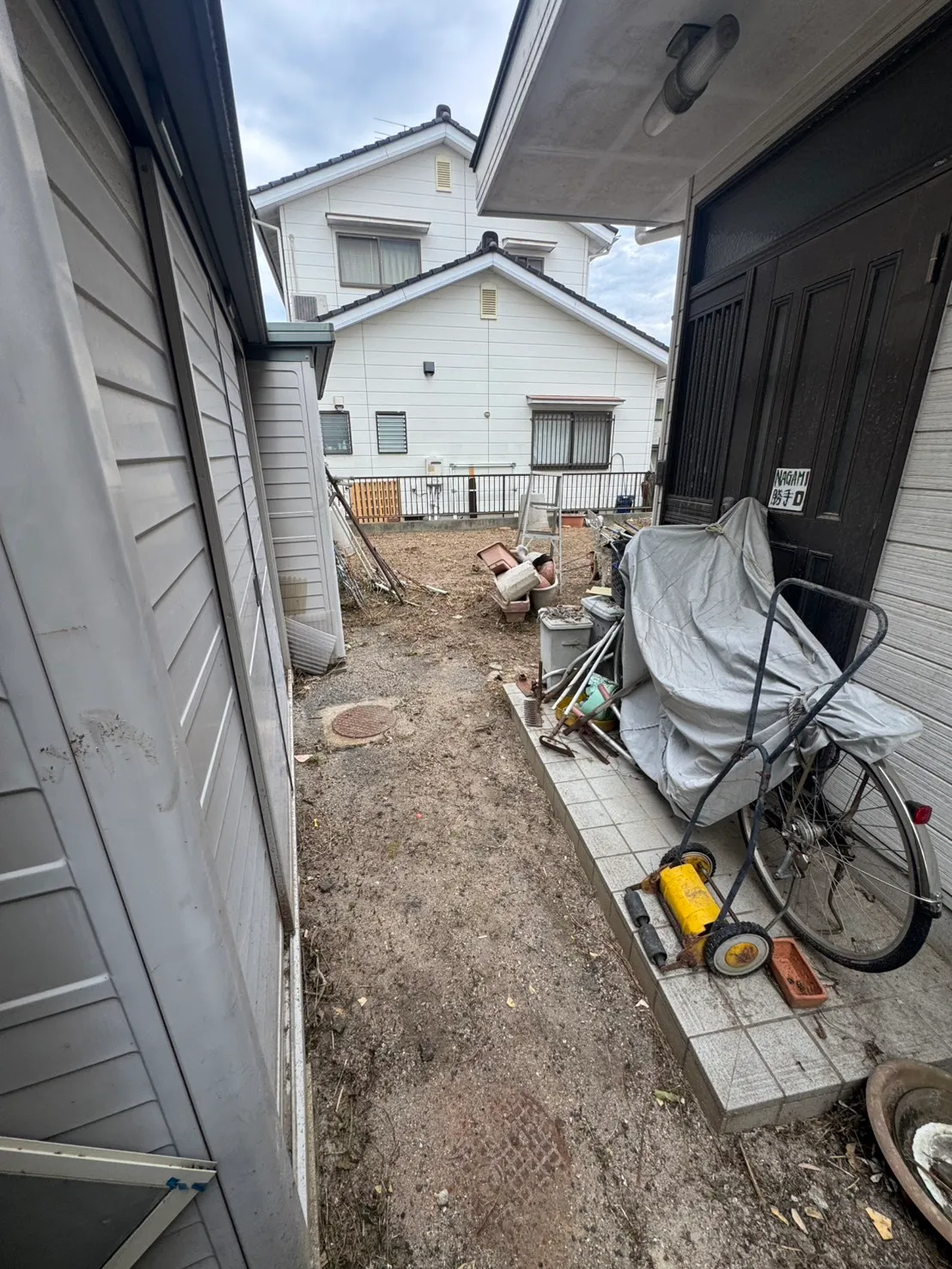 住宅の出入口と物置状の設備の間から庭へ続く地面。自転車や道具が脇に置かれている
